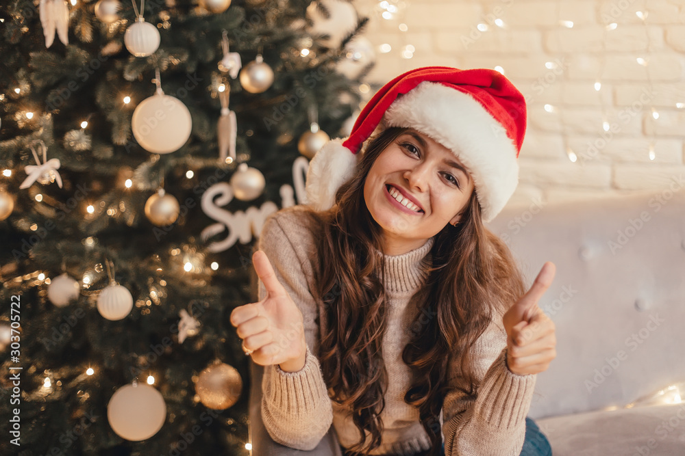 Woman in santa hat showing thumbs up sitting on the sofa near Christmas tree in the decorative interior. Positive gesture. Christmas and New Year photo.