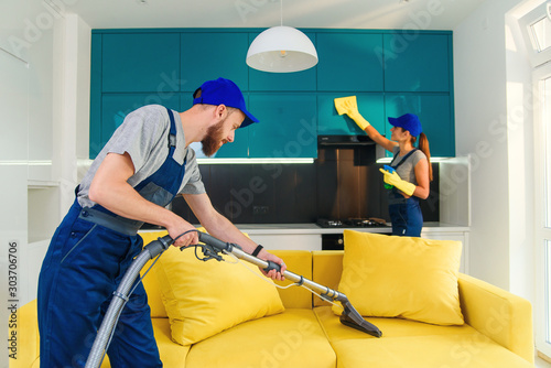 Young man vacuums yellow sofa and cute girl wiping kitchen furniture. Professional cleaners in apartment.