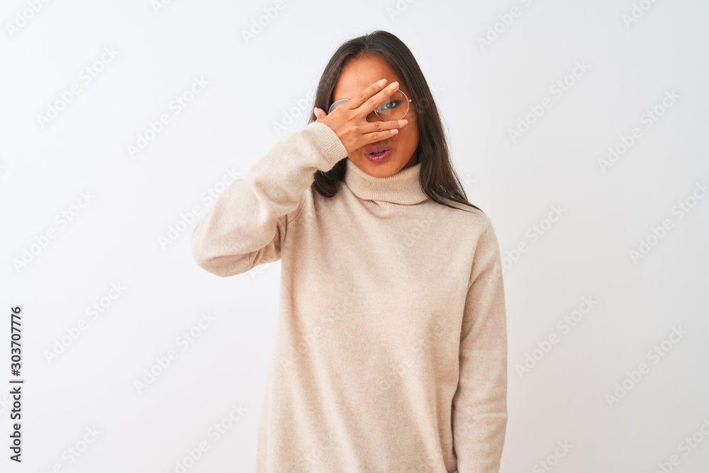 Young chinese woman wearing turtleneck sweater and glasses over isolated white background peeking in shock covering face and eyes with hand, looking through fingers with embarrassed expression. Young chinese woman wearing turtleneck sweater and glasses over isolated white background peeking in shock covering face and eyes with hand, looking through fingers with embarrassed expression.