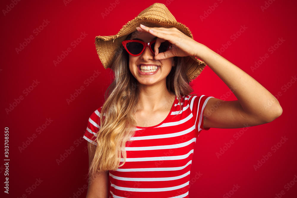 Young beautiful woman wearing sunglasses and summer hat over red isolated background doing ok gesture with hand smiling, eye looking through fingers with happy face.