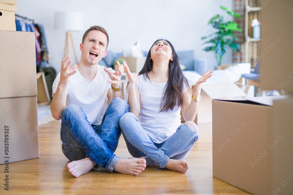 Young beautiful couple sitting on the floor at new home around cardboard boxes crazy and mad shouting and yelling with aggressive expression and arms raised. Frustration concept. Young beautiful couple sitting on the floor at new home around cardboard boxes crazy and mad shouting and yelling with aggressive expression and arms raised. Frustration concept.