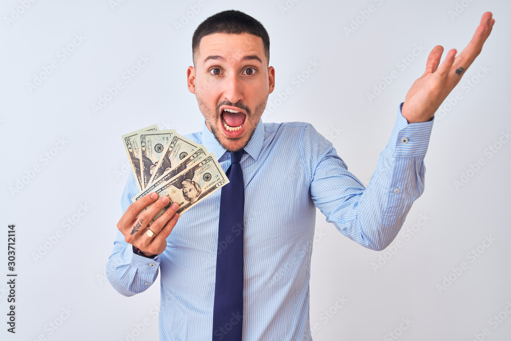 Young handsome business man holding a bunch of bank notes dollars over isolated background very happy and excited, winner expression celebrating victory screaming with big smile and raised hands Young handsome business man holding a bunch of bank notes dollars over isolated background very happy and excited, winner expression celebrating victory screaming with big smile and raised hands