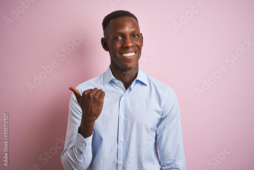 African american man wearing blue elegant shirt standing over isolated pink background smiling with happy face looking and pointing to the side with thumb up.