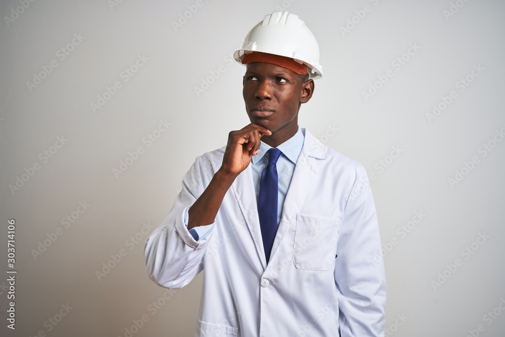 African american engineer man wearing coat and helmet over isolated white background with hand on chin thinking about question, pensive expression. Smiling with thoughtful face. Doubt concept. African american engineer man wearing coat and helmet over isolated white background with hand on chin thinking about question, pensive expression. Smiling with thoughtful face. Doubt concept.