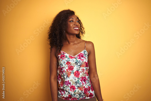 African american woman wearing floral summer t-shirt over isolated yellow background looking away to side with smile on face, natural expression. Laughing confident.