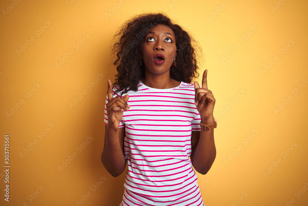 Young african american woman wearing striped t-shirt over isolated yellow background amazed and surprised looking up and pointing with fingers and raised arms. Young african american woman wearing striped t-shirt over isolated yellow background amazed and surprised looking up and pointing with fingers and raised arms.