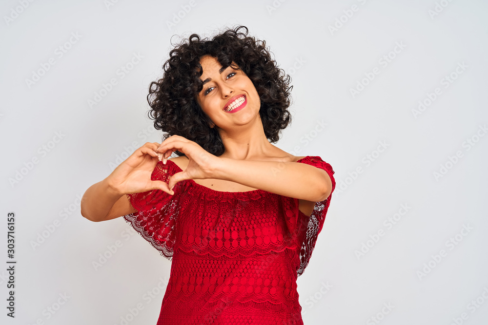 Young arab woman with curly hair wearing red dress standing over isolated white background smiling in love showing heart symbol and shape with hands. Romantic concept. Young arab woman with curly hair wearing red dress standing over isolated white background smiling in love showing heart symbol and shape with hands. Romantic concept.