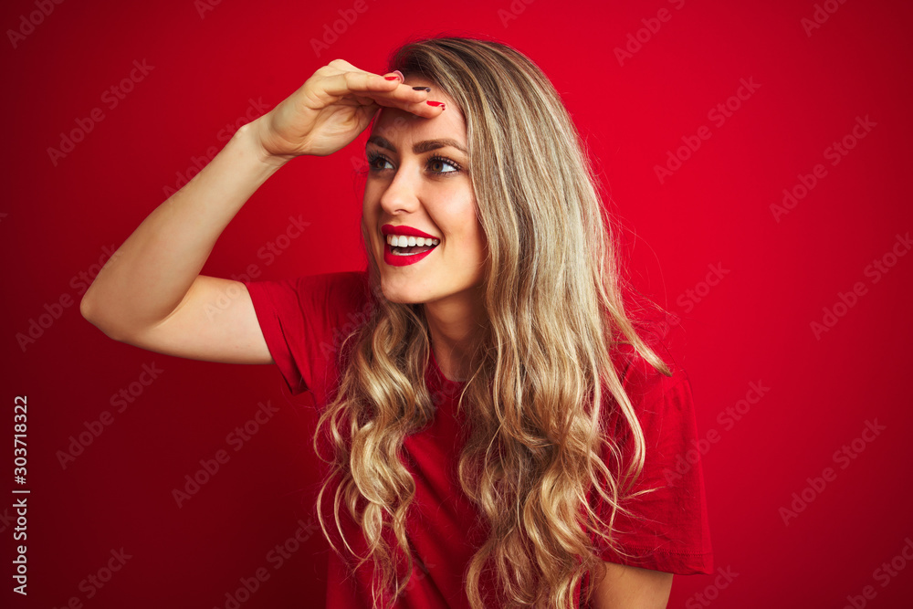 Young beautiful woman wearing basic t-shirt standing over red isolated background very happy and smiling looking far away with hand over head. Searching concept. Young beautiful woman wearing basic t-shirt standing over red isolated background very happy and smiling looking far away with hand over head. Searching concept.
