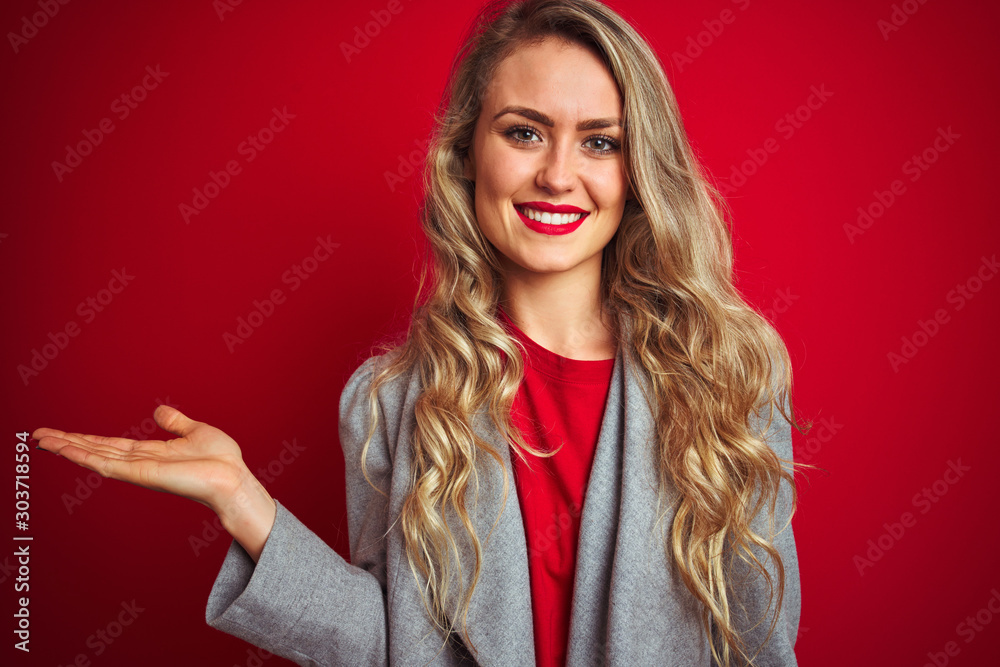 Young beautiful business woman wearing elegant jacket standing over red isolated background smiling cheerful presenting and pointing with palm of hand looking at the camera. Young beautiful business woman wearing elegant jacket standing over red isolated background smiling cheerful presenting and pointing with palm of hand looking at the camera.