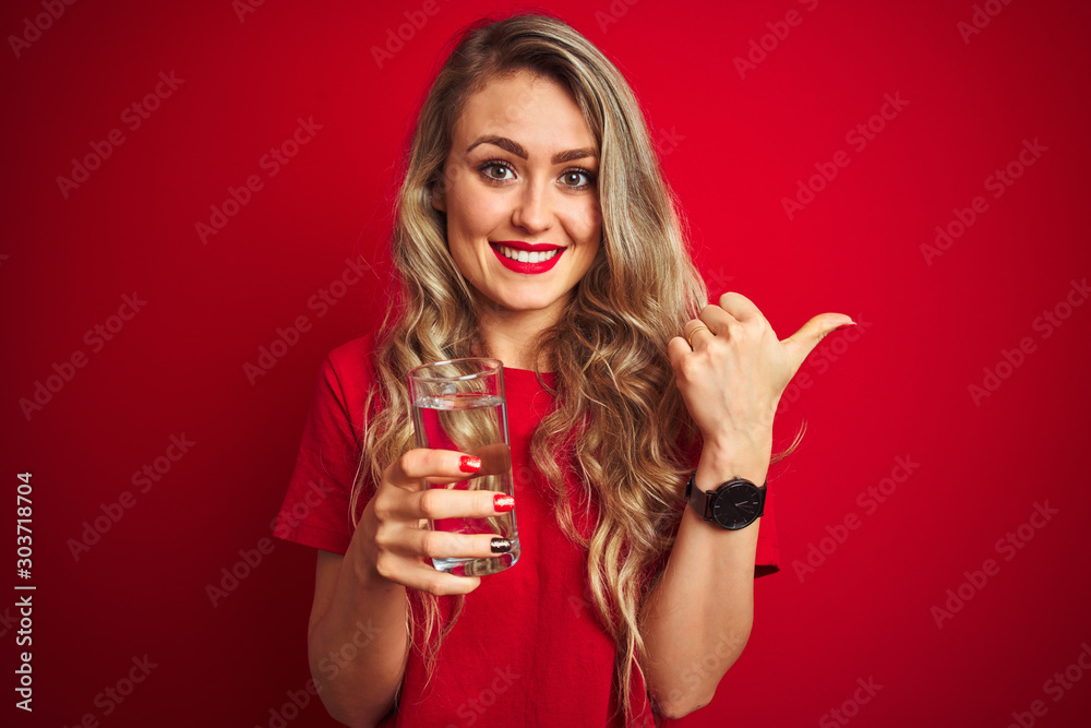 Young beautiful woman drinking a glass of water over red isolated background pointing and showing with thumb up to the side with happy face smiling Young beautiful woman drinking a glass of water over red isolated background pointing and showing with thumb up to the side with happy face smiling