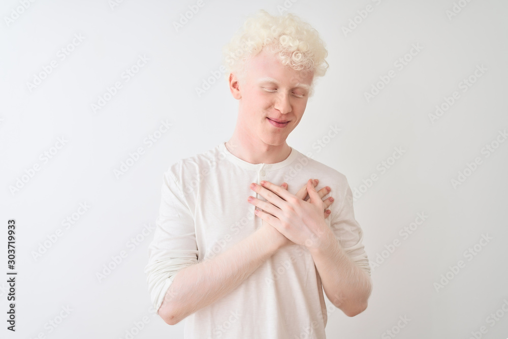 Young albino blond man wearing casual t-shirt standing over isolated white background smiling with hands on chest with closed eyes and grateful gesture on face. Health concept. Young albino blond man wearing casual t-shirt standing over isolated white background smiling with hands on chest with closed eyes and grateful gesture on face. Health concept.
