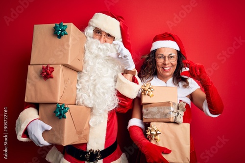 Middle age couple wearing Santa costume holding tower of gifts over isolated red background smiling and confident gesturing with hand doing small size sign with fingers looking and the camera. 