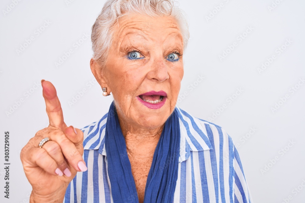 Senior grey-haired woman wearing blue striped shirt standing over isolated white background surprised with an idea or question pointing finger with happy face, number one Senior grey-haired woman wearing blue striped shirt standing over isolated white background surprised with an idea or question pointing finger with happy face, number one