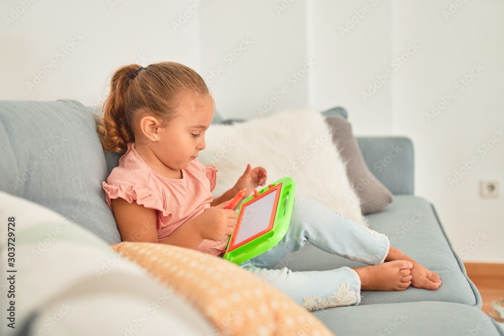 Beautiful blond toddler girl sitting on the sofa drawing using digital board at home Beautiful blond toddler girl sitting on the sofa drawing using digital board at home