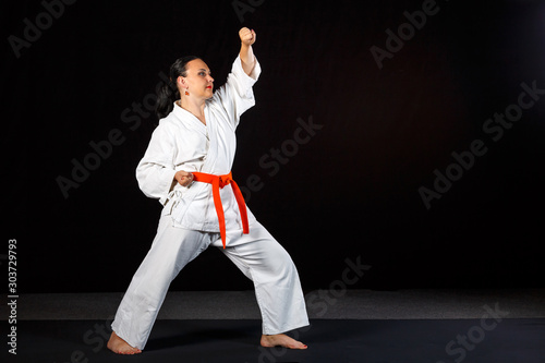 A young brunette woman in a white kimono doing karate on a black background in full growth.