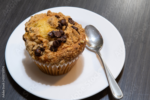 Big chocolate chip muffin or cupcake on white plate with small metal spoon on plate. Gray table background. Breakfast, desert concept.