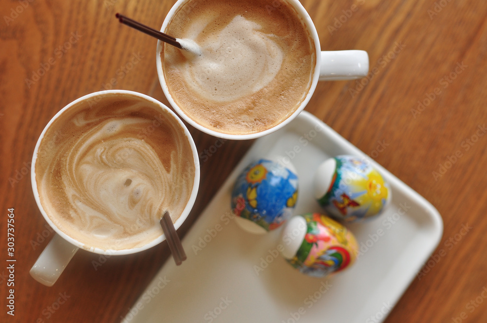 Happy Easter holiday in a cafe, two white cups of coffee on a wooden table, a white plate with colored eggs Happy Easter holiday in a cafe, two white cups of coffee on a wooden table, a white plate with colored eggs