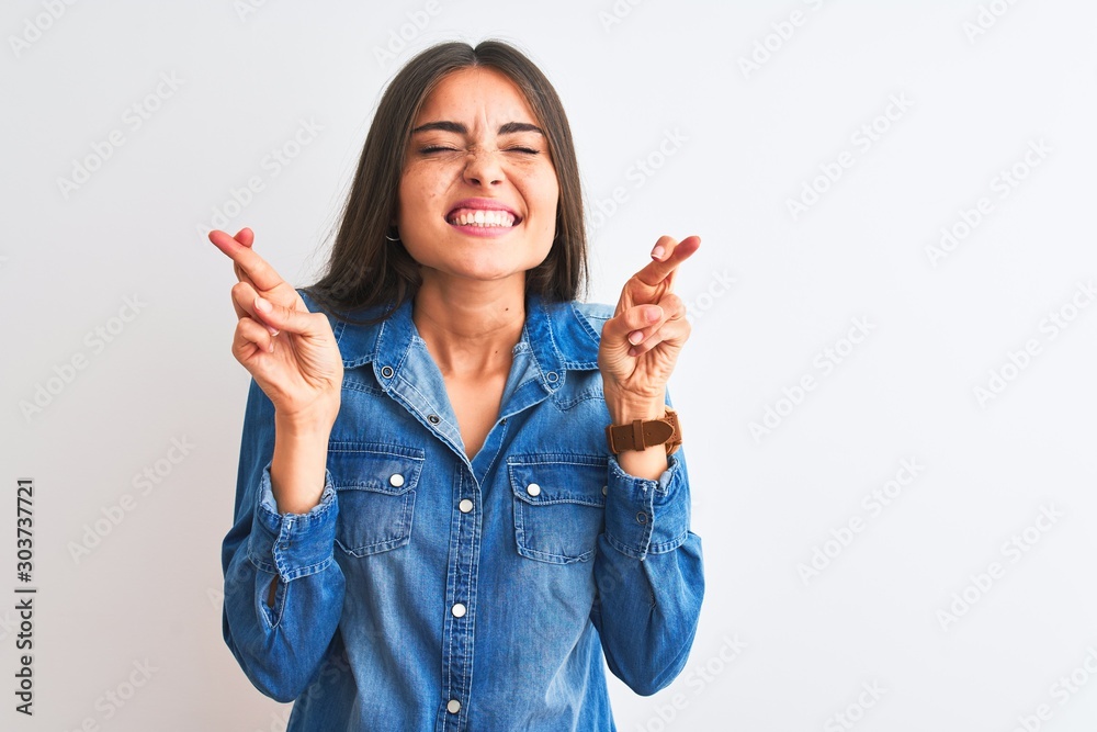 Young beautiful woman wearing casual denim shirt standing over isolated white background gesturing finger crossed smiling with hope and eyes closed. Luck and superstitious concept. Young beautiful woman wearing casual denim shirt standing over isolated white background gesturing finger crossed smiling with hope and eyes closed. Luck and superstitious concept.