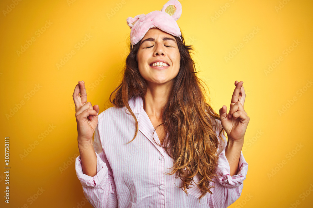 Φωτογραφία Young woman wearing pajama and sleep mask standing over yellow isolated background gesturing finger crossed smiling with hope and eyes closed