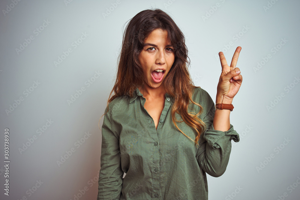 Φωτογραφία Young beautiful woman wearing green shirt standing over grey isolated background smiling with happy face winking at the camera doing victory sign with fingers