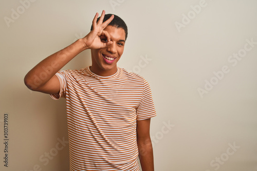 Young handsome arab man wearing striped t-shirt over isolated white background doing ok gesture with hand smiling, eye looking through fingers with happy face. © Krakenimages.com