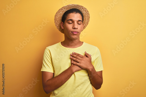 Young handsome arab man wearing t-shirt and summer hat over isolated yelllow background smiling with hands on chest with closed eyes and grateful gesture on face. Health concept.