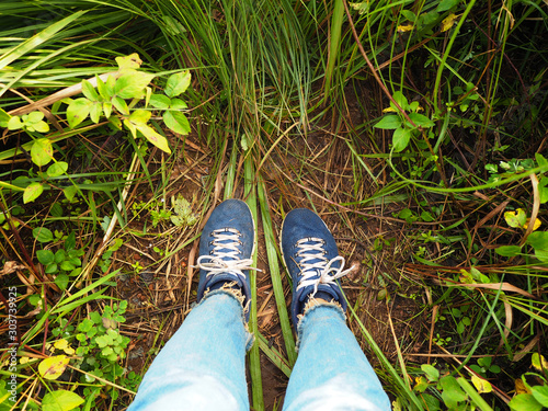 Woman selfie feet wearing dirty sneakers and blue jeans on green wild grass.