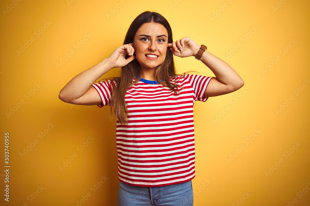 Young beautiful woman wearing striped t-shirt standing over isolated yellow background covering ears with fingers with annoyed expression for the noise of loud music. Deaf concept. Young beautiful woman wearing striped t-shirt standing over isolated yellow background covering ears with fingers with annoyed expression for the noise of loud music. Deaf concept.