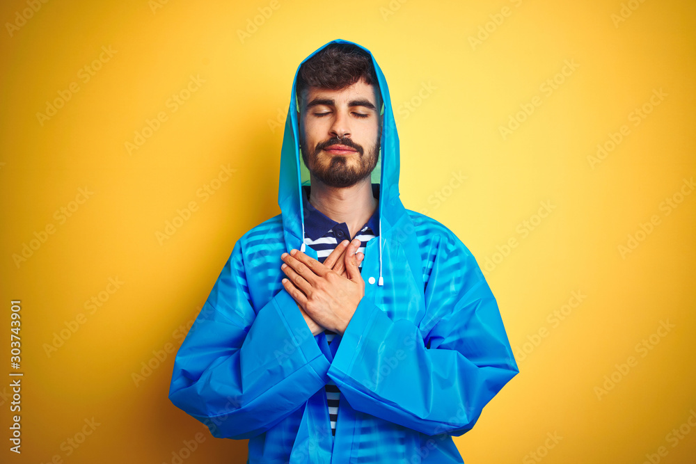 Young handsome man wearing rain coat with hood standing over isolated yellow background smiling with hands on chest with closed eyes and grateful gesture on face. Health concept. Young handsome man wearing rain coat with hood standing over isolated yellow background smiling with hands on chest with closed eyes and grateful gesture on face. Health concept.