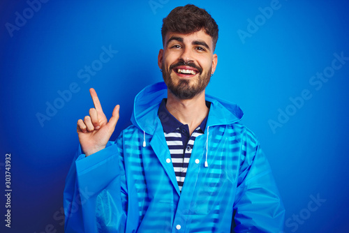 Young handsome man wearing rain coat standing over isolated blue background with a big smile on face, pointing with hand and finger to the side looking at the camera.