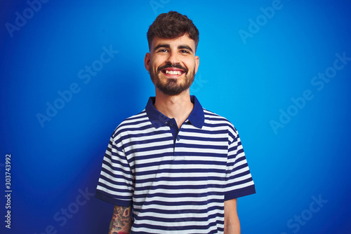 Young man with tattoo wearing striped polo standing over isolated blue background with a happy and cool smile on face. Lucky person.