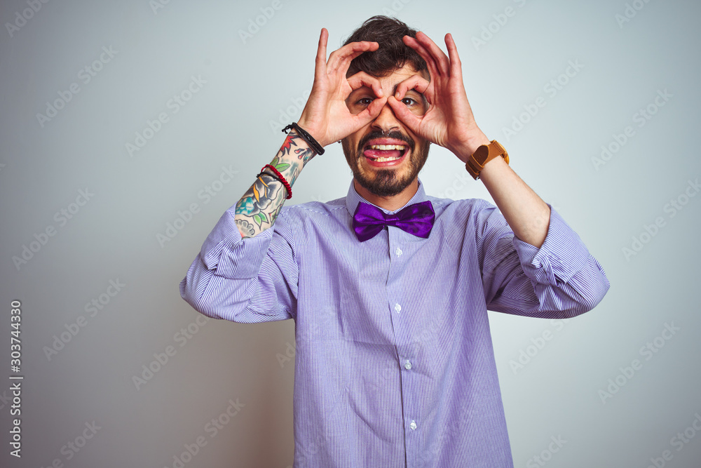 Young man with tattoo wearing purple shirt and bow tie over isolated white background doing ok gesture like binoculars sticking tongue out, eyes looking through fingers. Crazy expression. Young man with tattoo wearing purple shirt and bow tie over isolated white background doing ok gesture like binoculars sticking tongue out, eyes looking through fingers. Crazy expression.