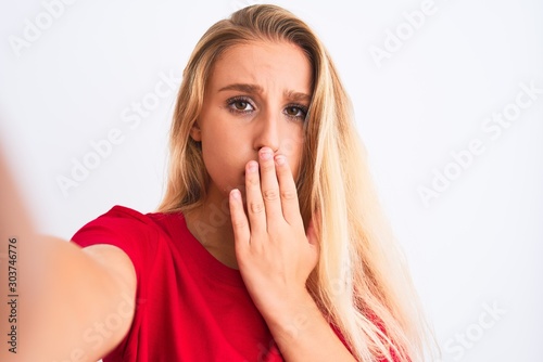 Young beautiful woman wearing t-shirt make selfie by camera over isolated white background cover mouth with hand shocked with shame for mistake, expression of fear, scared in silence, secret concept