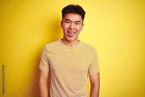 Young asian chinese man wearing t-shirt standing over isolated yellow background sticking tongue out happy with funny expression. Emotion concept.