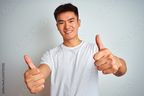 Young asian chinese man wearing t-shirt standing over isolated white background approving doing positive gesture with hand, thumbs up smiling and happy for success. Winner gesture. © Krakenimages.com