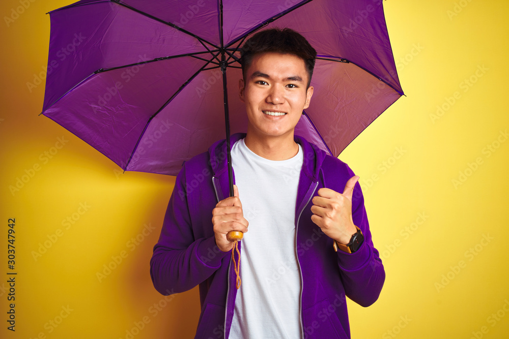 Εκτύπωση καμβά Asian chinese man wearing purple umbrella standing over isolated yellow backgrou