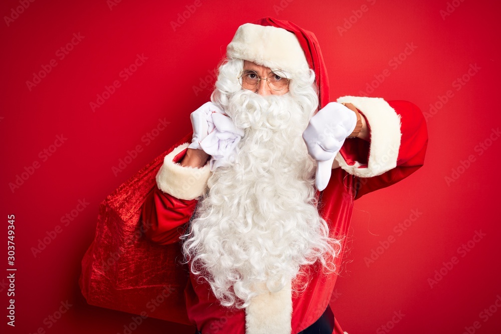 Middle age man wearing Santa costume holding sack with gifts over isolated red background with angry face, negative sign showing dislike with thumbs down, rejection concept