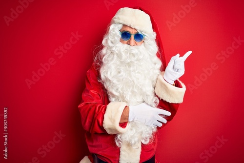 Middle age man wearing Santa Claus costume and sunglasses over isolated red background with a big smile on face, pointing with hand and finger to the side looking at the camera.