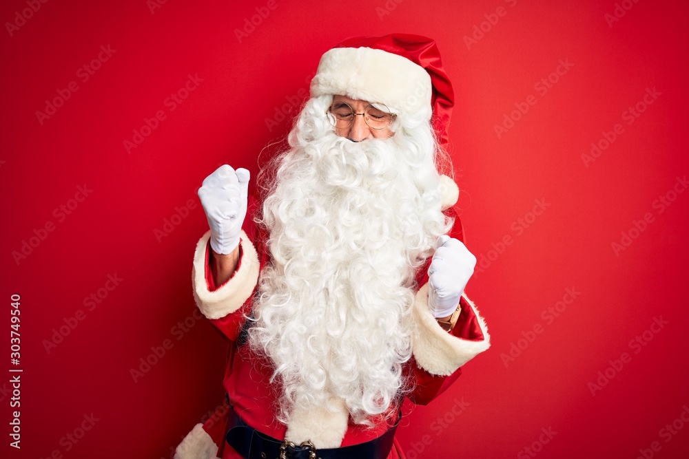 Middle age handsome man wearing Santa costume standing over isolated red background very happy and excited doing winner gesture with arms raised, smiling and screaming for success. Celebration concept