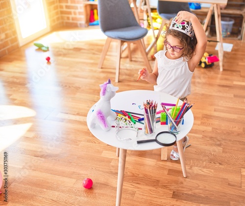 Beautiful toddler wearing glasses and princess crown sitting drawing using paper and marker pen at kindergarten