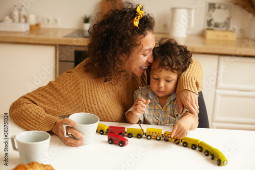 Beautiful young Hispanic mom embracing adorable baby boy, kissing him on forehead, having morning coffee, sitting at kitchen table, playing with toys together in the morning. Family and generations photo