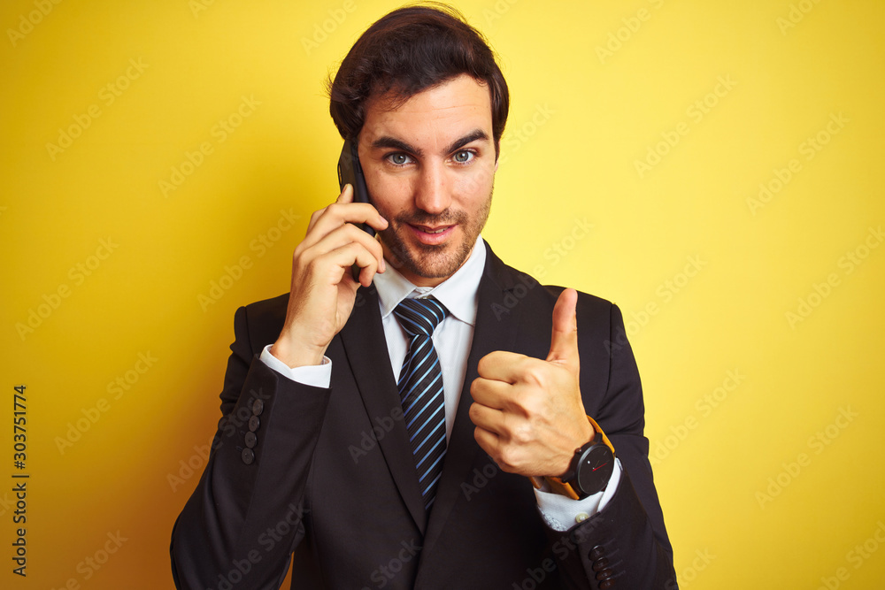 Young handsome businessman talking on the smartphone over isolated yellow background happy with big smile doing ok sign, thumb up with fingers, excellent sign Young handsome businessman talking on the smartphone over isolated yellow background happy with big smile doing ok sign, thumb up with fingers, excellent sign