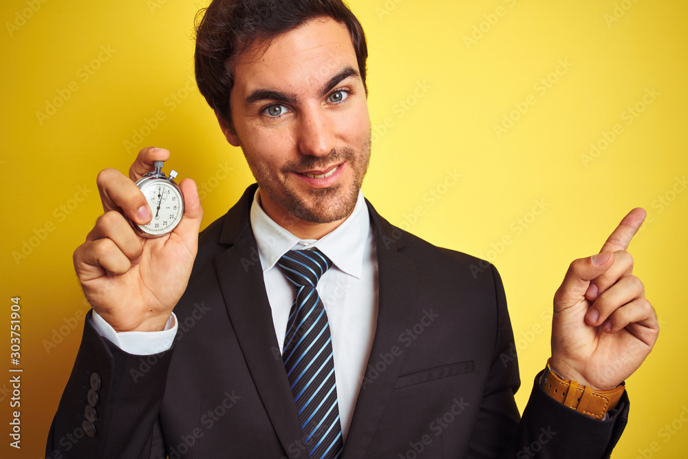 Young handsome businessman holding stopwatch standing over isolated yellow background very happy pointing with hand and finger to the side Young handsome businessman holding stopwatch standing over isolated yellow background very happy pointing with hand and finger to the side