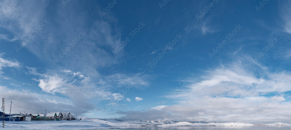 View of russian countryside landscape in winter sunny day with freezing lake and picturesque cloudy blue sky reflecting on water with new transcparent ice surface; scenic village in Ural mountains