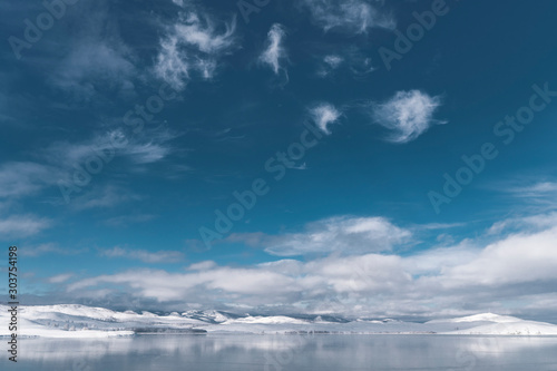 Panorama of winter landscape near Ural mountains; sunny snowy day with deep blue cloudy sky on freezing lake with hills reflection on water surface; season changing in wild nature, Russia
