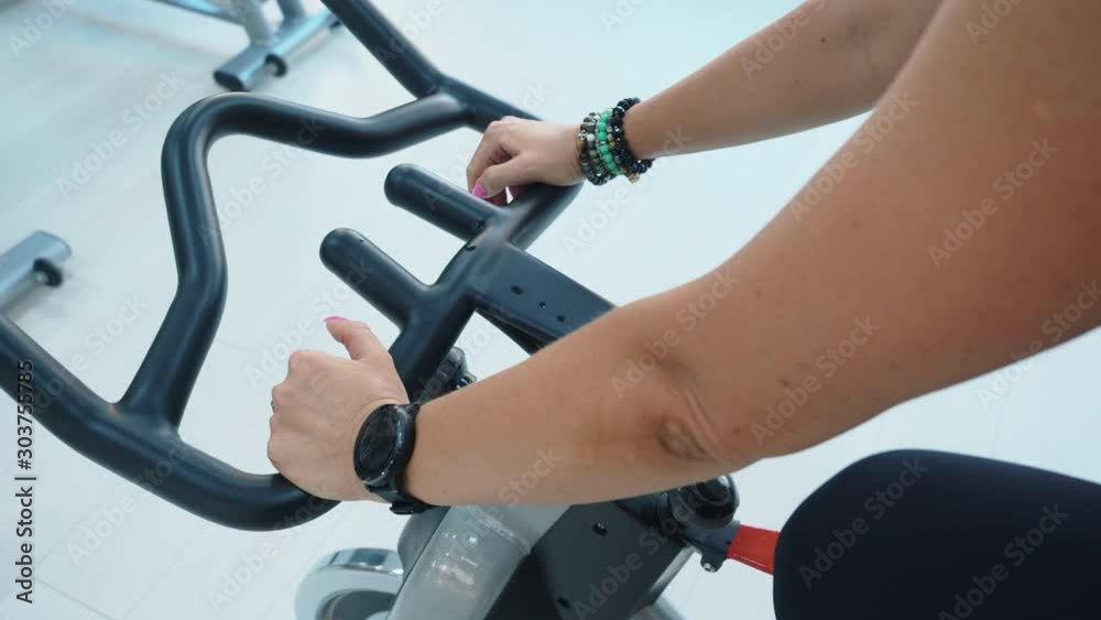 Female hands with fitness watch holding handlebar of indoor bike at cycling class in sport club. Slim woman talk and gestures training on static bike in gym. Fitness woman spinning bike machine Female hands with fitness watch holding handlebar of indoor bike at cycling class in sport club. Slim woman talk and gestures training on static bike in gym. Fitness woman spinning bike machine