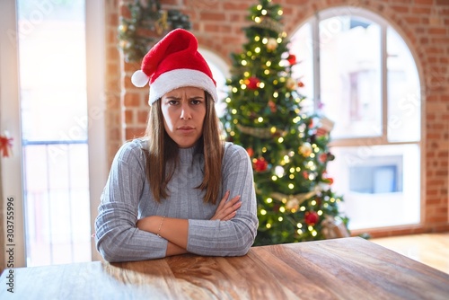 Young beautiful woman wearing santa claus hat at the table at home around christmas decoration skeptic and nervous, disapproving expression on face with crossed arms. Negative person.