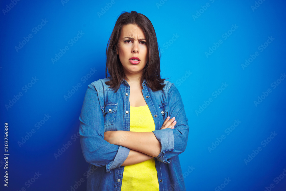 Young beautiful brunette woman wearing casual blue denim jacket over blue isolated background skeptic and nervous, disapproving expression on face with crossed arms. Negative person. Young beautiful brunette woman wearing casual blue denim jacket over blue isolated background skeptic and nervous, disapproving expression on face with crossed arms. Negative person.