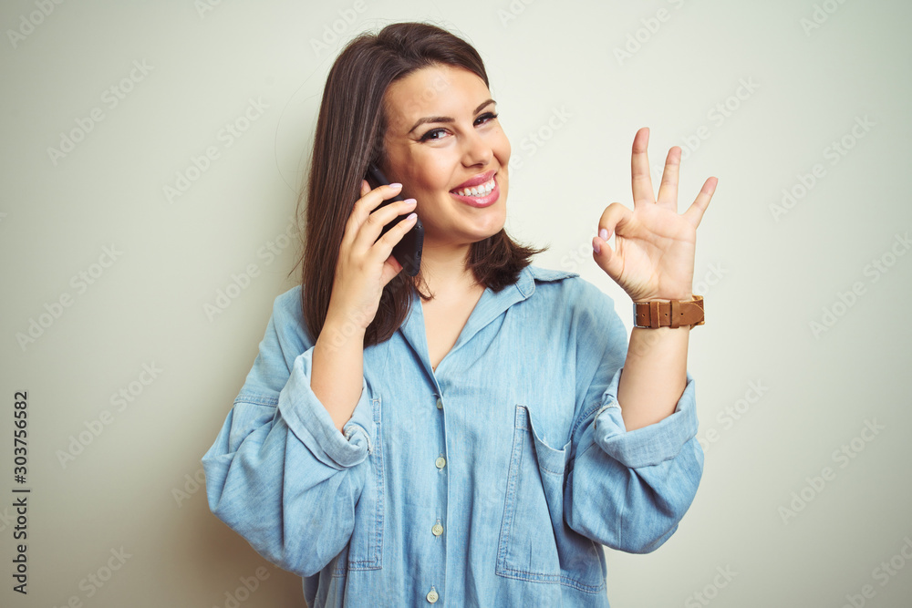 Young beautiful woman having a conversation using smartphone over isolated background doing ok sign with fingers, excellent symbol Young beautiful woman having a conversation using smartphone over isolated background doing ok sign with fingers, excellent symbol