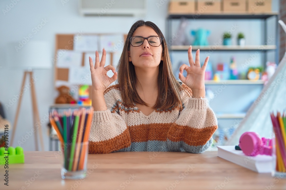 Φωτογραφία Young beautiful teacher woman wearing sweater and glasses sitting on desk at kindergarten relaxed and smiling with eyes closed doing meditation gesture with fingers